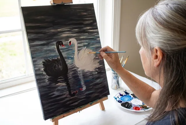 A woman at an easel painting the black swan and white swan scene, with a palette of black, white, red, and blue paint beside her