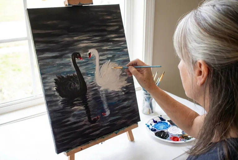 A woman at an easel painting the black swan and white swan scene, with a palette of black, white, red, and blue paint beside her
