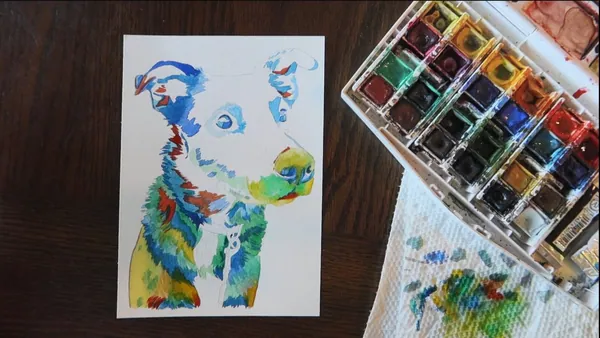 A part-finished rainbow watercolor dog portrait beside a well-used watercolor palette